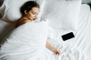 https://www.pexels.com/photo/a-woman-sleeping-beside-her-smartphone-9787924/ Alt Text: Woman asleep and smiling in bed with her phone next to her